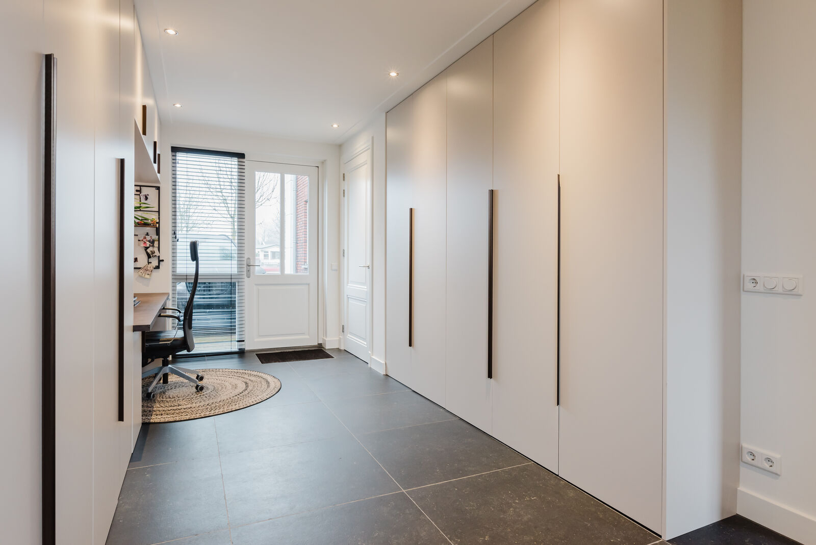 Kastenwand op maat over de volledige lengte van een hal, met verticale bronskleurige greeplijsten en aan het einde een werkplek op maat bij het raam