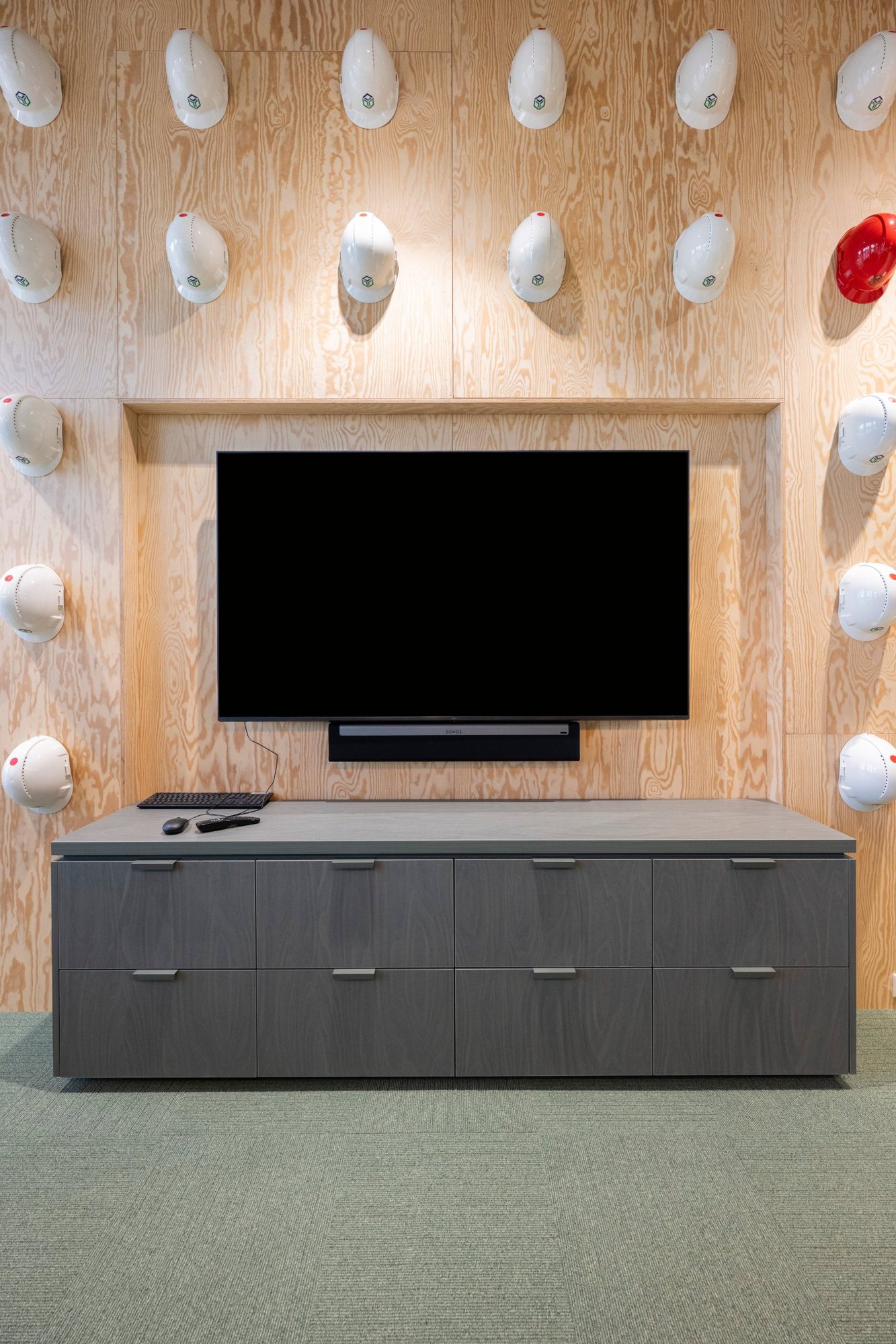 Houten wand op maat met witte bouwhelmen, één rode helm, tv nis en grijs dressoir in kantoor Bouwmij Janssen