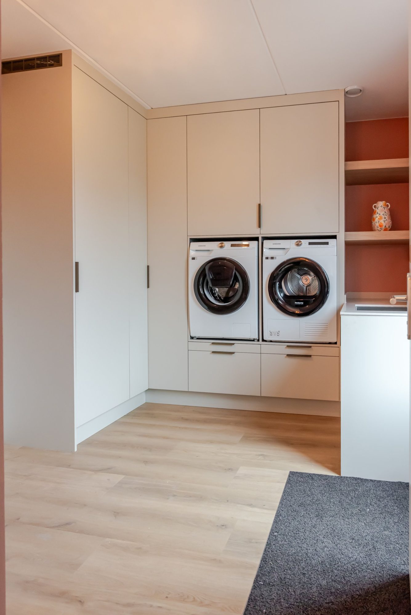 Waskamer op maat met plafondhoge kasten, wasmachine en droger op hoogte en eiken planken in een terracotta nis