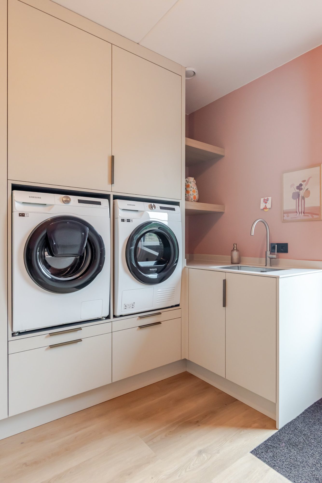 Wasmachine en droger naast elkaar op hoogte in een kast op maat, met spoelbak en roze wand